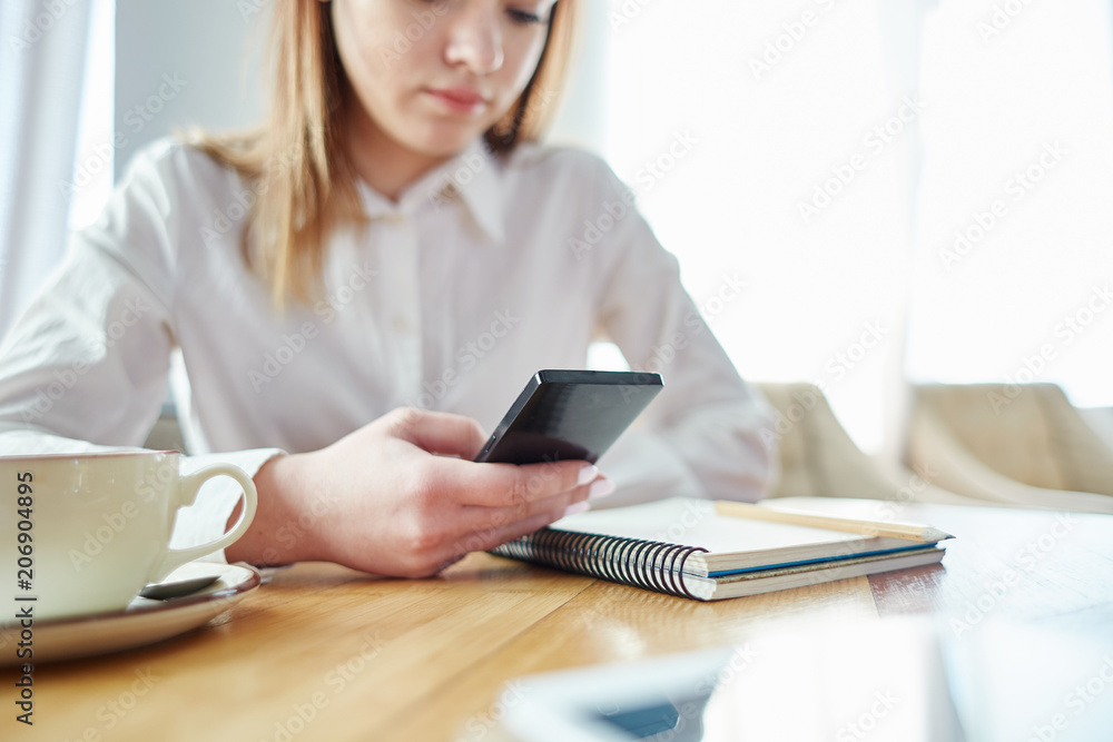 Young businesswoman sitting in cafe and texting on cell phone; coffee cup, notebook and pencil on table. Focus on hand holding smartphone Young businesswoman sitting in cafe and texting on cell phone; coffee cup, notebook and pencil on table. Focus on hand holding smartphone