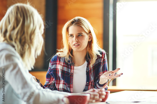 Two young female friends chatting over coffee in cafe. Blonde women discussing issues