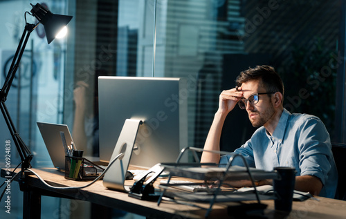 Foto Casual tired office worker sitting at desk using computer and doing overtime project in lamplight