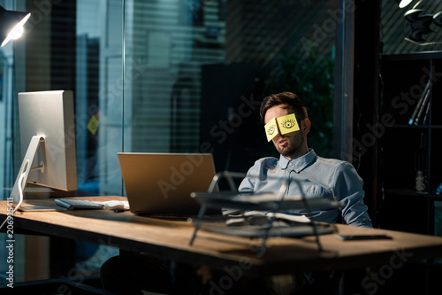 Office worker sleeping at table covering eyes with stickers having painted eyes.  photo