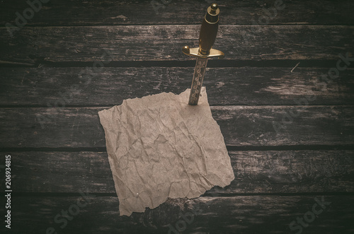 Fotografie Crumpled brown paper page parchment and the dagger blade thrust in a table