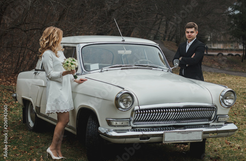 Just married wedding couple is standing near the retro vintage car in the park. Spring or autumn grey day in forest. Blonde bride in elegant white dress with bouquet and elegant groom in love. © Vadym