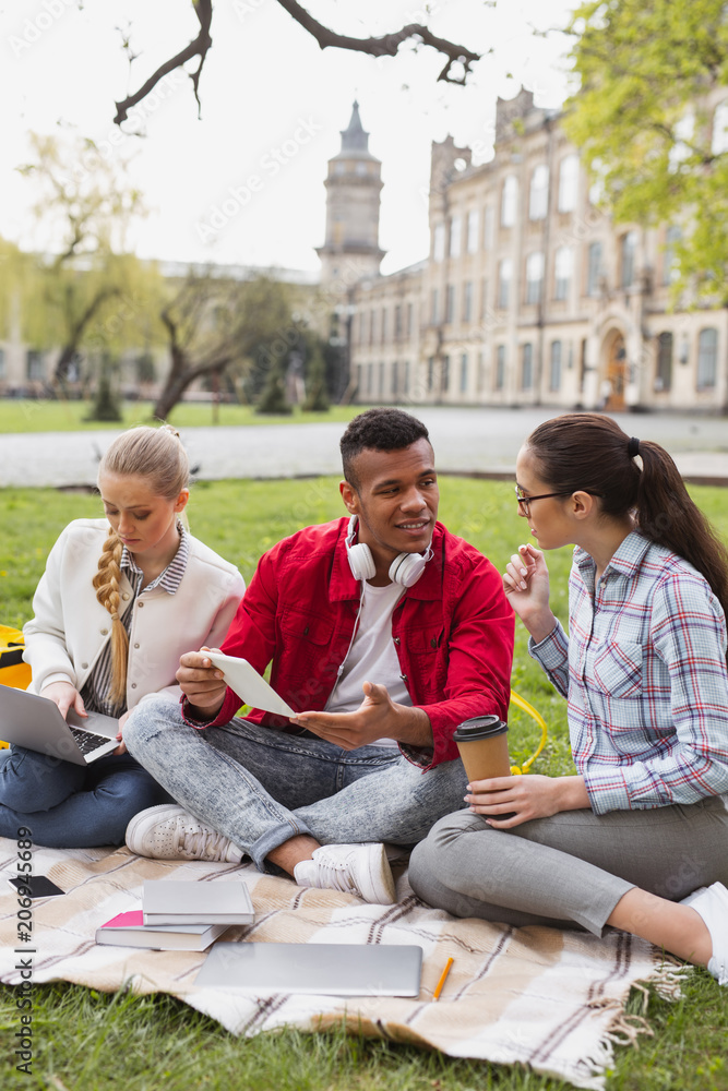 Young cute couple feeling amazing while sitting near their beautiful groupmate spending weekend in the park Young cute couple feeling amazing while sitting near their beautiful groupmate spending weekend in the park