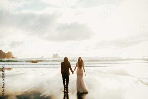 Adorable wedding couple holds each other hands walking along the beach against the rocks and ocean waves in the evening light photo