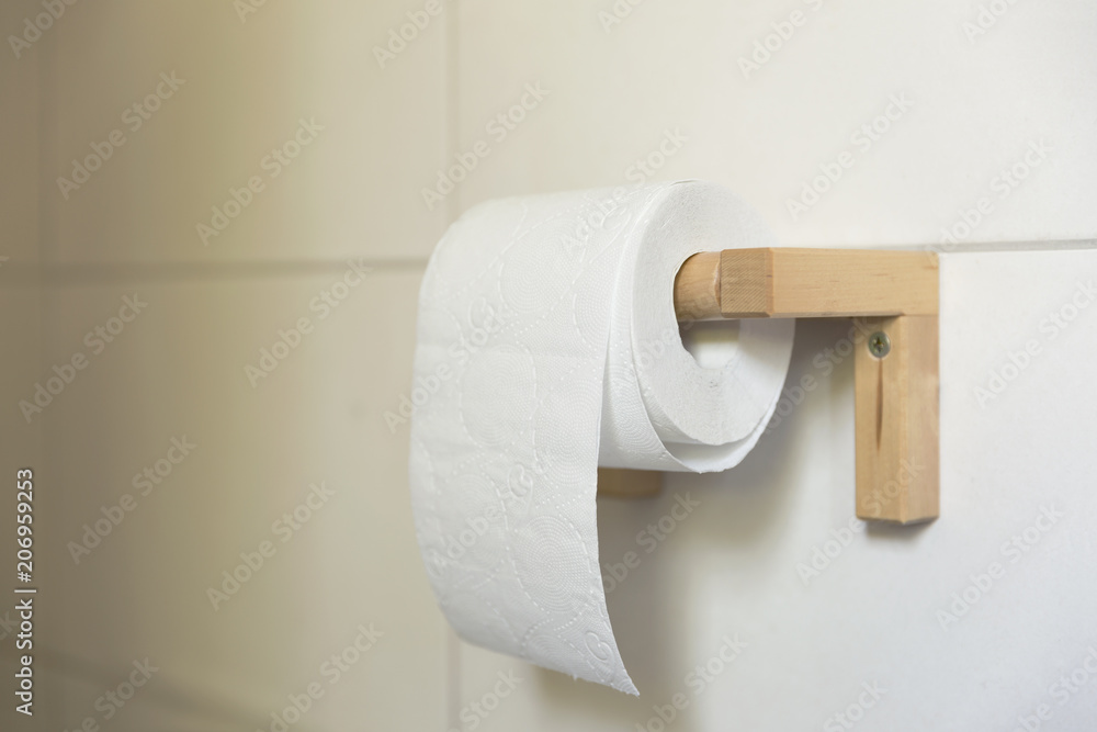 Obraz premium A white roll of soft toilet paper neatly hanging on a modern wooden holder on a white tile bathroom wall.