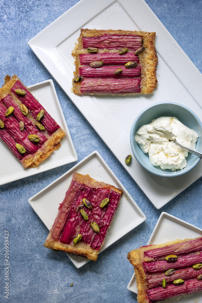Servings of rhubarb tart sprinkled with pistachio nuts on dessert plates with a bowl of cream. Servings of rhubarb tart sprinkled with pistachio nuts on dessert plates with a bowl of cream.