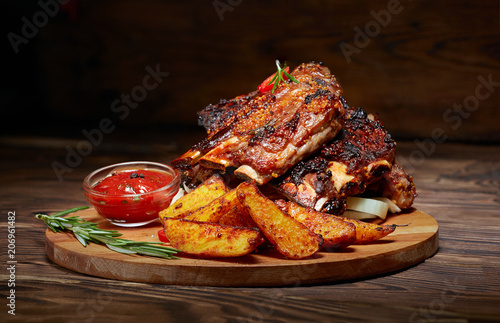 Fried ribs with rosemary, potatoes rustic, onion, sauce on wooden round Board. Dark background. Place for text, copyspace
