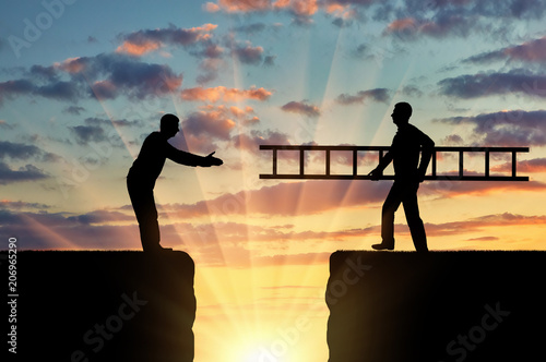 A silhouette of a man carries a ladder to another man who is on the other side of the abyss photo
