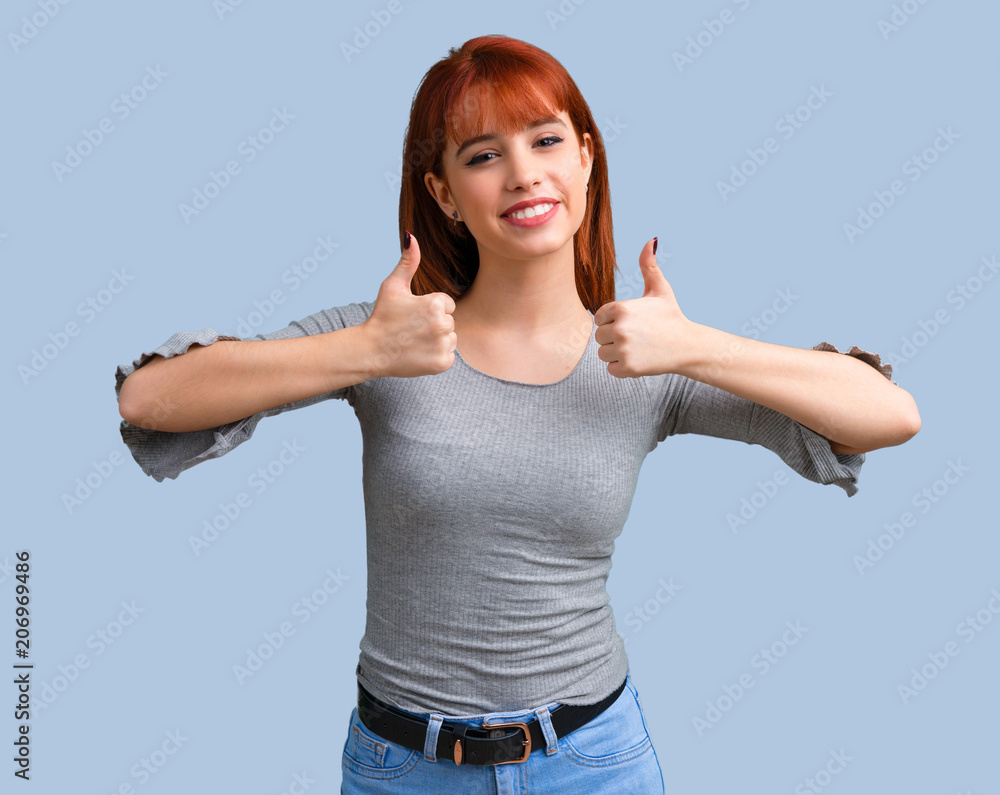 Young redhead girl giving a thumbs up gesture and smiling on blue background Young redhead girl giving a thumbs up gesture and smiling on blue background