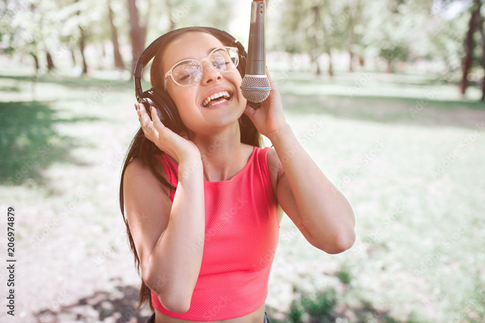 Portrait of girl standing outside and singing to the mic. She is listening to music at the same time while she is singing. Young woman is enjoying the moment. Portrait of girl standing outside and singing to the mic. She is listening to music at the same time while she is singing. Young woman is enjoying the moment.