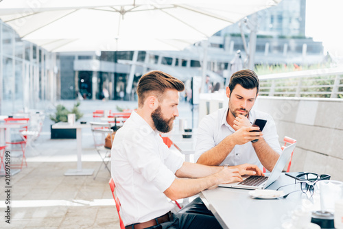 two bearded young business young men outdoor using laptop and smart phone remote working - finance, trading online, wifi technology concept