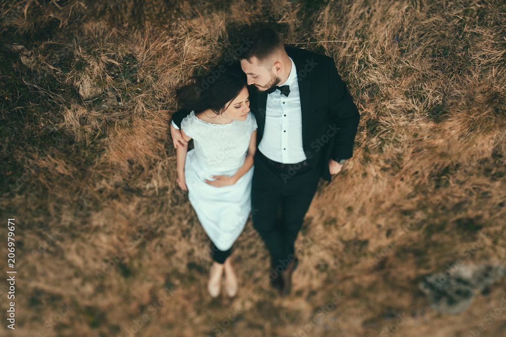Fototapeta premium Top view shot of beautiful sensual wedding couple of bride and groom lying and hugging on the grass in the mountains.