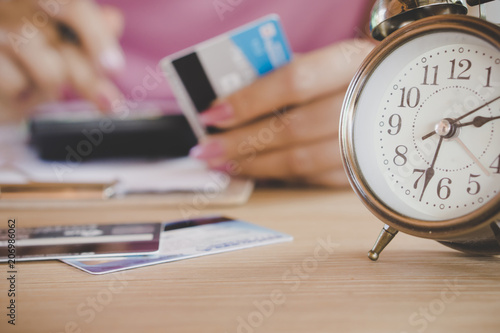 blur background of woman hand calculating her expenses with some credit cards and alarm clock on desk 