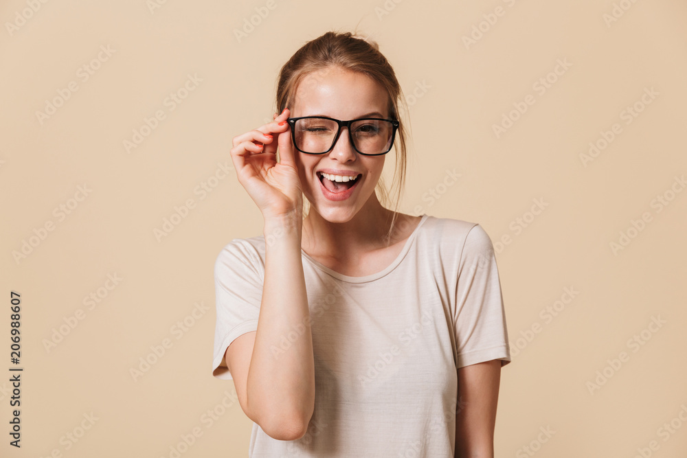 Portrait of beautiful cheerful woman 20s wearing basic t-shirt touching eyeglasses and smiling at camera while winking, isolated over beige background in studio