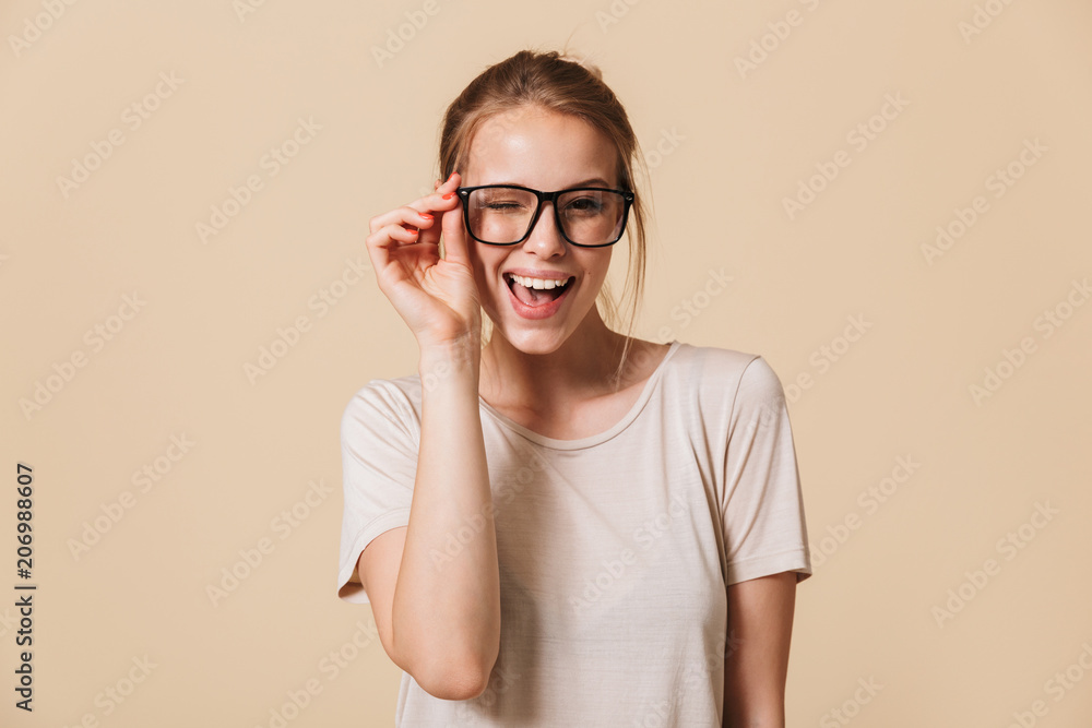 Portrait of beautiful cheerful woman 20s wearing basic t-shirt touching eyeglasses and smiling at camera while winking, isolated over beige background in studio Portrait of beautiful cheerful woman 20s wearing basic t-shirt touching eyeglasses and smiling at camera while winking, isolated over beige background in studio
