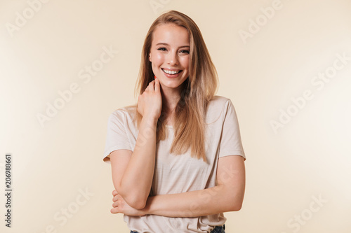 Portrait of charming blonde woman 20s wearing casual t-shirt smiling at camera while touching neck, isolated over beige background in studio