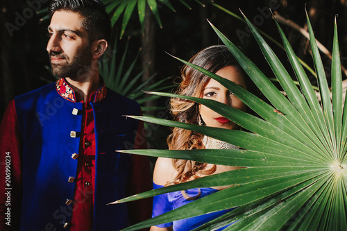 Gorgeous Hindu groom in blue and red sherwani and bride in blue lehenga pose in the dark garden photo