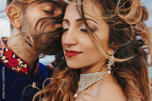 Wind blows around Hindu groom in blue sherwani and bride in lehenga posing in white house with gorgeous seaside view behind them photo