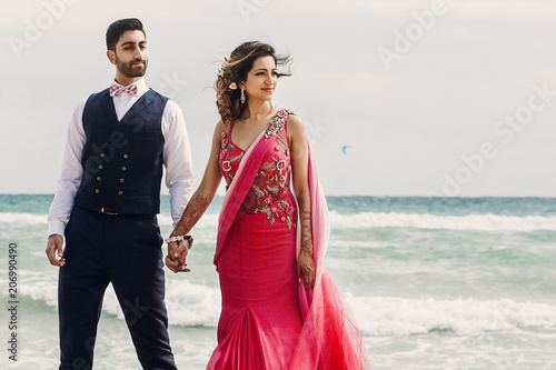 Wind blows bride's pink sari while she stands with Hindu groom on a wooden quay among foaming waves photo