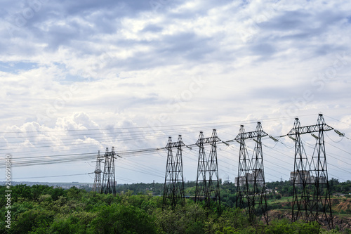 High voltage lines and power pylons in a green forest, sunny day with clouds in the sky. in the background building, city, village, power supply, electrification, electricity transmission problems