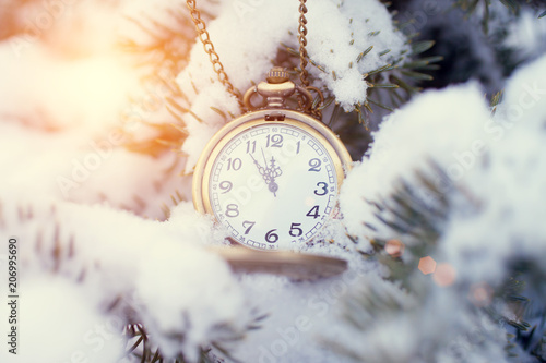 the clock on the chain hangs on the branches of the Christmas tree in the snow. The time is indicated at 11:55 photo