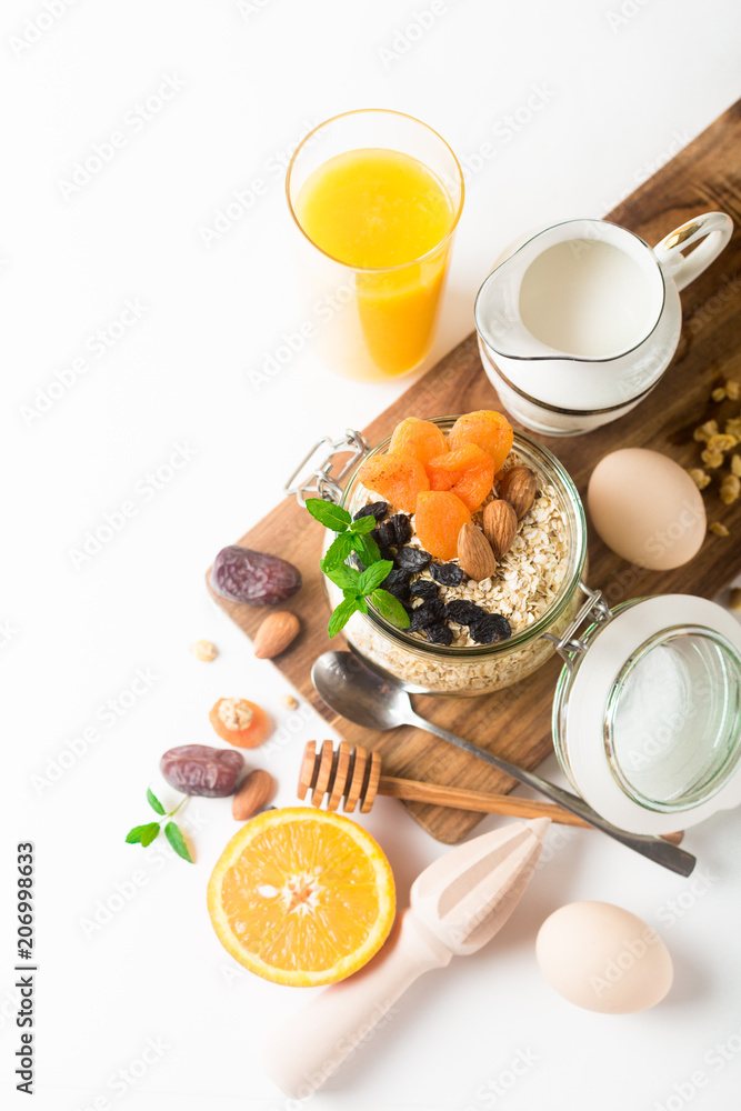 Healthy breakfast in glass mason jar with oat flakes, granola, milk, dry fruits and nuts, dates, honey, orange juice, eggs for morning fitness on wooden board platter on white background isolated Healthy breakfast in glass mason jar with oat flakes, granola, milk, dry fruits and nuts, dates, honey, orange juice, eggs for morning fitness on wooden board platter on white background isolated
