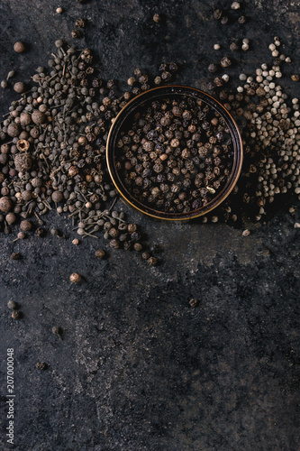 Variety of different black peppers allspice, pimento, monks pepper, peppercorns in tin can over old black iron texture background. Top view, copy space.