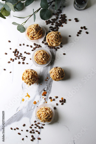 Fresh baked homemade cupcakes with coffee buttercream and caramel with eucalyptus branch and coffee beans above over white marble background. Flat lay, space