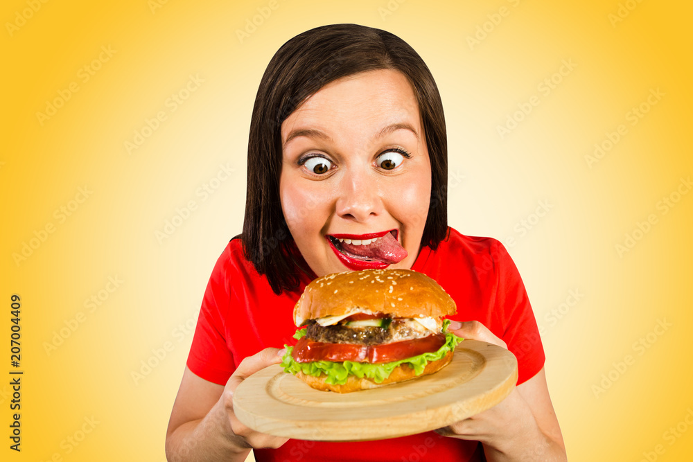 Young woman holds hamburger, smiles, looks on him and licks. Isolated on orange background.