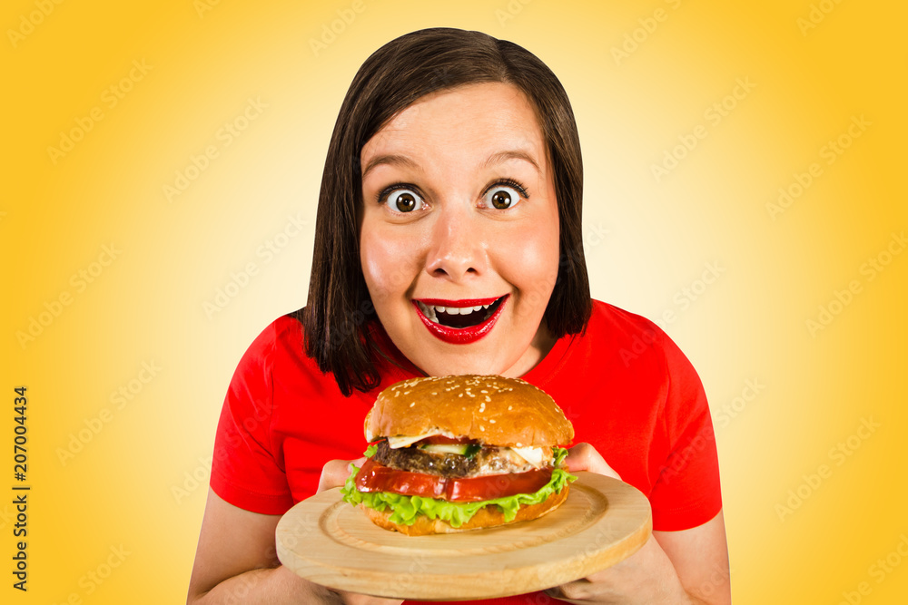 Young woman holds hamburger, smiles and looks in camera. Isolated on orange background.