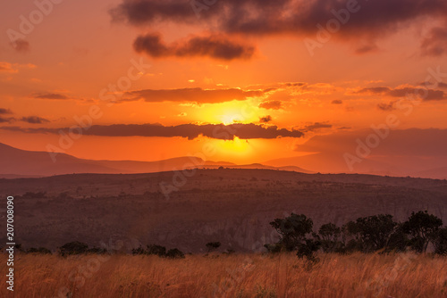 The sun glows behind clouds in an intense sunset above an African landscape of hills, cliffside and savannah at Blyde River Canyon in Mpumalanga, South Africa