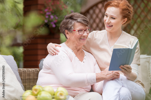 Cheerful senior woman with her tender caretaker reading a book together while relaxing outside. Close relationship.