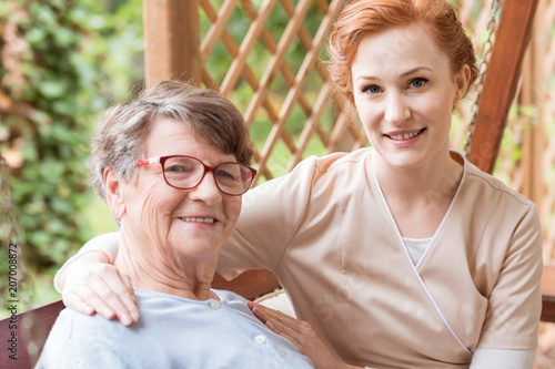 Bilde på lerret Close-up of a professional caregiver sitting next to and holding her arm around a senior lady on a patio outside of a family home