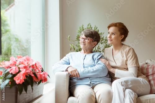 Картината върху платно Geriatric woman and her professional caretaker sitting on a couch and looking through a window in a living room