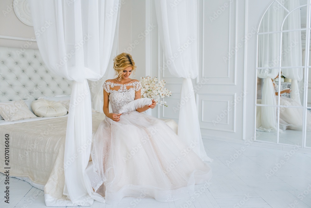 Fototapeta premium A bride with hairstyle and make up looking at the mirror preparing for the wedding. A portrait of beautiful girl with blond hair and blue eyes in wedding dress in studio with flowers