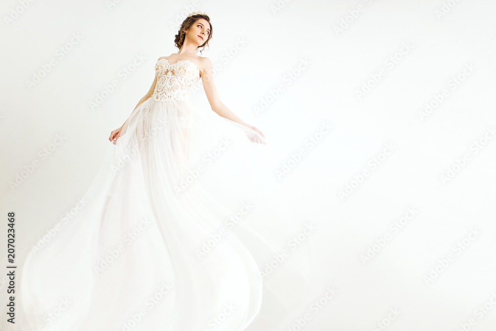 young smily brunette sexy girl in a white wedding boudoir dress and crown in hairstyle is dancing  like swan princess in motion  on a white wall background 