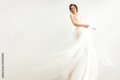 young smily brunette sexy girl in a white wedding boudoir dress and crown in hairstyle is dancing like swan princess in motion on a white wall background 