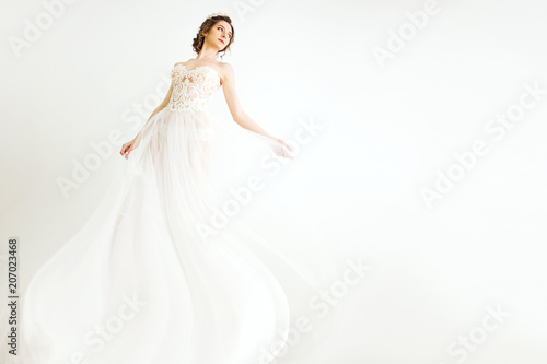 young smily brunette sexy girl in a white wedding boudoir dress and crown in hairstyle is dancing  like swan princess in motion  on a white wall background 