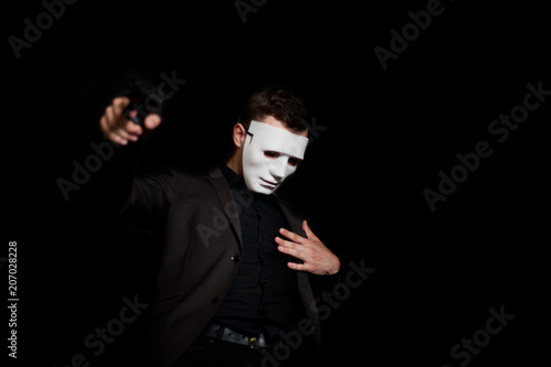 A man in a white mask on a black background, holding a gun and touching the wound with his hand