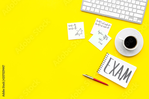 Preparing for the exam. Word Exam written in notebook on student's desk with computer, cup of coffee, cheat sheets on yellow backgrond top view space for text