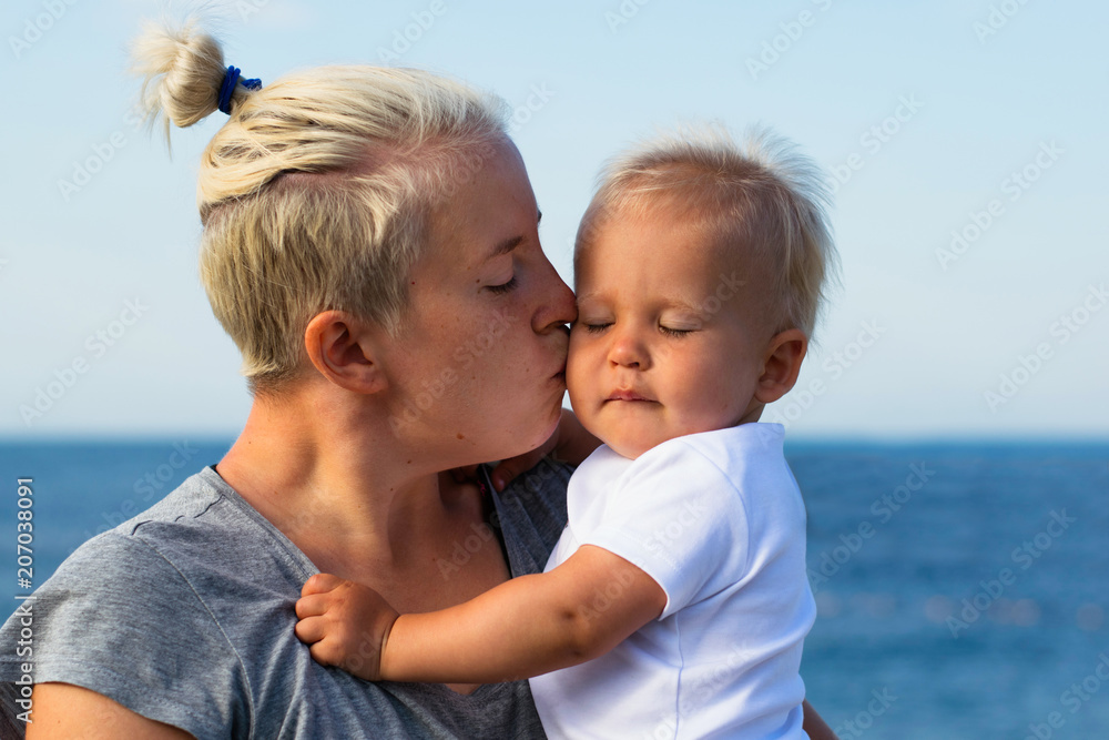 Fototapeta premium Young hipster mother kisses her blonde baby with closed eyes. Portrait of mom and child against blue sea. Motherhood, mother and baby relationship and tenderness concept