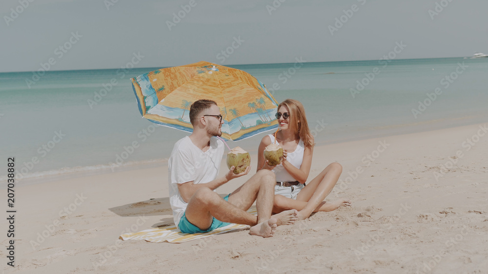 Young happy couple drinking coconut milk on the tropical beach during sunny summer holiday