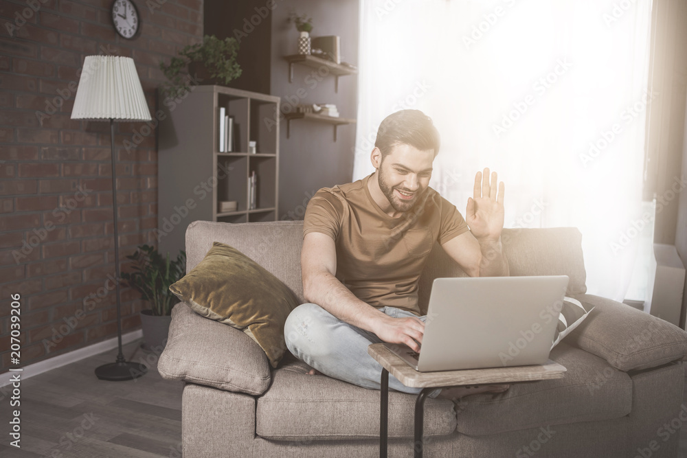 Hi there. Full-length portrait of cheerful guy is sitting on cozy sofa and enjoying video call on his modern laptop. He is emotionally waving his hand while talking in net. Sunlight in background Hi there. Full-length portrait of cheerful guy is sitting on cozy sofa and enjoying video call on his modern laptop. He is emotionally waving his hand while talking in net. Sunlight in background