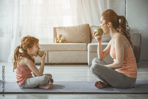 Profile of woman and kid after aerobics. They are sitting in front of each other on the floor and biting green apples with smile