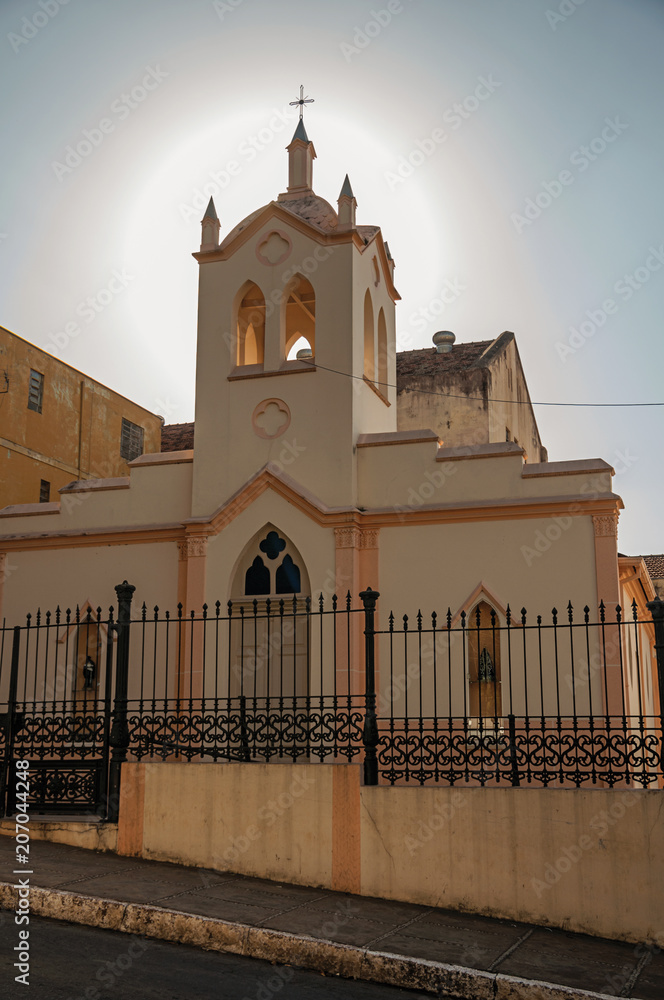 Facade of small church and belfry, behind iron fence, with sunshine behind at sunset in São Manuel. A cute little town in the countryside of São Paulo State. Southeast Brazil.