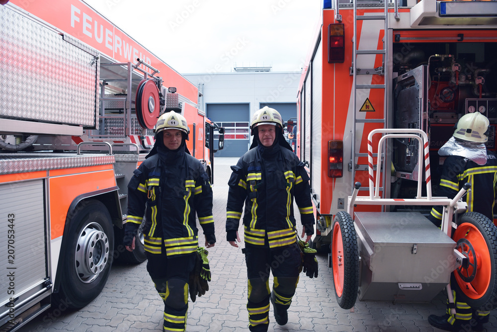 Naklejka premium Feuerwehrmänner am Löschfahrzeug in Schutzausrüstung // Firemen at the vehicle in front of a fire station