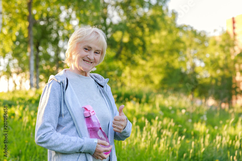 Happy elderly woman shows thumbs up in sportswear in a sunny park. Be active and energetic until old age, longevity, biohacking