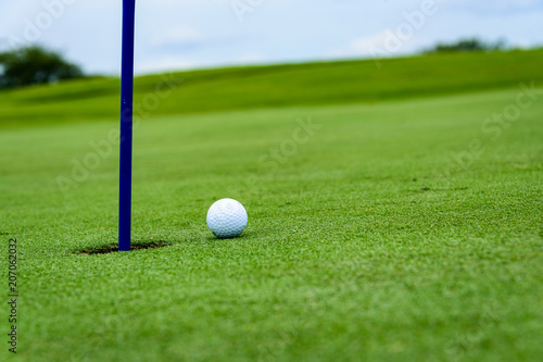 Side view of a golf ball on a sloped putting green, close to the hole with a blue pin, with sky, clouds, and bushes in the background 