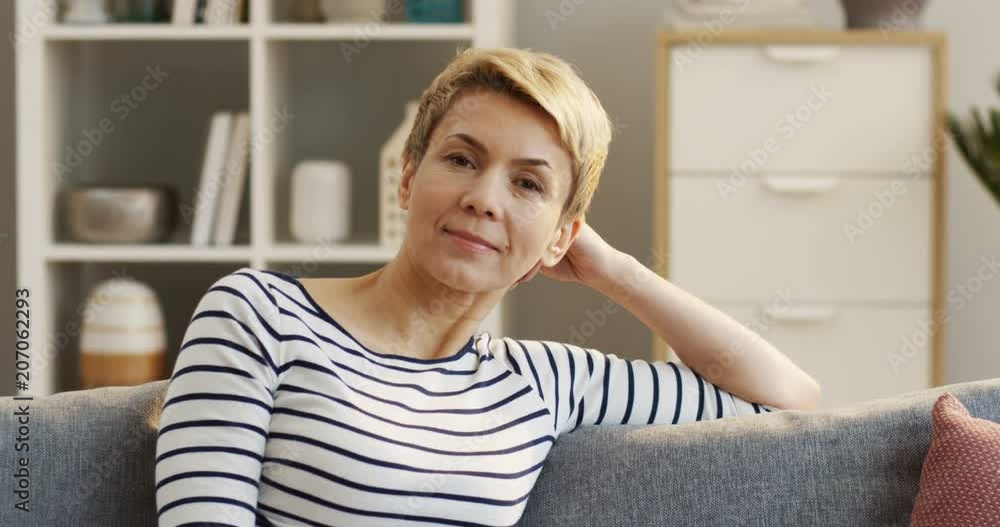 Portrait of the beautiful woman of middle aged woman in the striped blouse sitting on the gray couch and leaning on the hand. Indoors Portrait of the beautiful woman of middle aged woman in the striped blouse sitting on the gray couch and leaning on the hand. Indoors