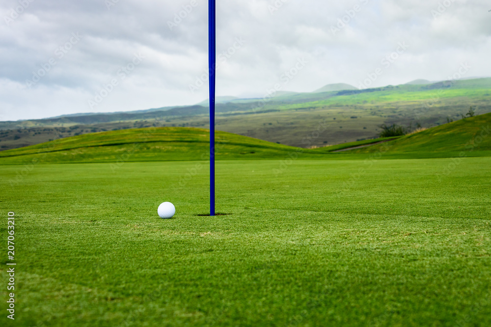 Obraz premium Close up side view of a golf ball on a putting green next to the hole with a blue pin, and hills and a stormy sky in the background